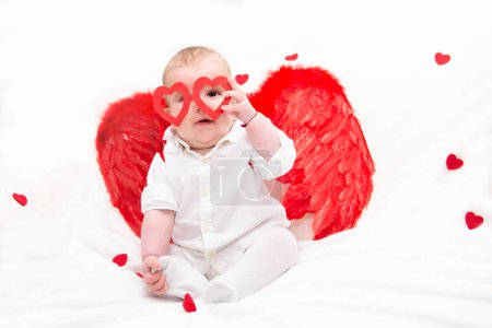 Téléchargez les photos : Mignon enfant mignon, tient des lunettes en forme de coeur avec un stylo sur un fond blanc avec des coeurs avec des ailes d'ange rouge.Félicitations pour la Saint-Valentin. Cupidon gamin - en image libre de droit