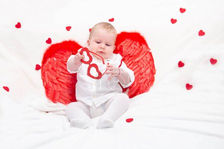 Téléchargez les photos : Un bébé aux joues bouffies en vêtements blancs avec des ailes de Cupidon rouge, joue avec des lunettes avec des coeurs, isolé sur une couverture blanche tendre avec des coeurs. Cupidon enfant pour un projet avec les amoureux. - en image libre de droit