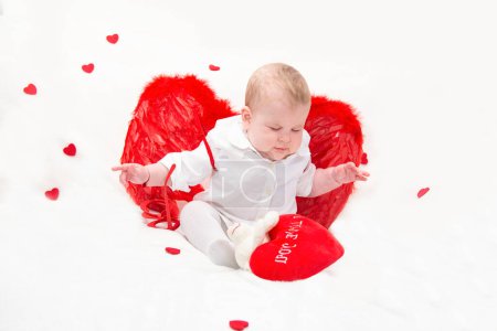 Téléchargez les photos : Un bébé avec des joues gonflées en vêtements blancs avec des ailes de Cupidon rouge avec les yeux fermés, assis avec les bras écartés, mouches, isolé sur un fond blanc avec des coeurs - en image libre de droit