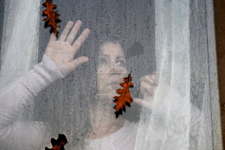Téléchargez les photos : Une femme regarde tristement les feuilles orange qui tombent à travers une fenêtre mouillée avec des gouttes de pluie.L'homme regarde la rue à travers la fenêtre, profitant du temps d'automne à l'extérieur. concept de temps pluvieux d'automne. - en image libre de droit