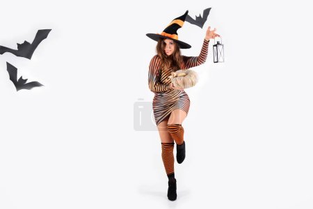 Frau mit Hexenhut und Halloween-Kleidung mit Laterne und Kürbis in der Hand, lachend, aufspringend, posierend, in die Kamera blickend mit schwarzen Fledermäusen auf weißem Hintergrund