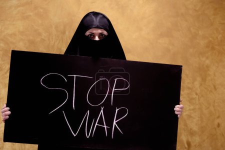 Photo for Palestinian woman in a hijab with a tired,sad look holding a banner with the inscription in chalk calling to stop the war against the abstract backdrop. - Royalty Free Image