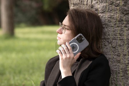 Photo for Guy with glasses in a business jacket and jeans,holding an Apple iPhone 14 PRO MAX to his ear,talking and listening against the background of nature in the park. - Royalty Free Image