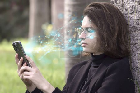Photo for A guy wearing glasses and business clothes holds a cell phone, looks at the screen with outgoing waves of holograms of information and neural connections transmitted to the brain. The concept of the Negative impact of AI on humans through Telecommuni - Royalty Free Image