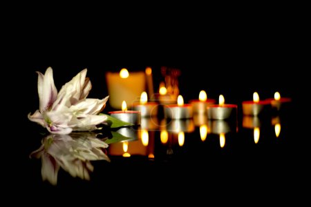 White lily and burning candles In the silence of the darkness of a funeral on a black plaque with the reflection of lights. Sad mourning background