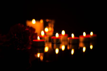 Sad mourning blurred background of burning candles, carnation flower In the silence of the darkness of a funeral on a black memorial plaque with reflection