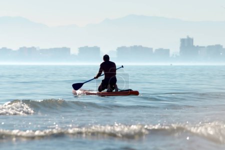 Silhouette of a kayaker on a personal rowing kayak, rowing a kayak with an oar in the waters of the ocean at dawn against the background of city buildings. Summer sports holiday on the water