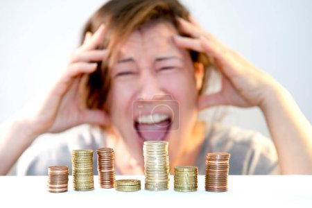 A man holds his head with his hands, screams with a grimace of horror, anxiety and helplessness, stress from lack of money in front of stacks of coins. Economic problems, financial crisis