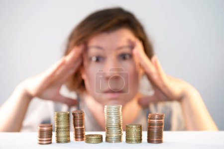 A man holds his head in his hands, looks at stacks of coins with sadness, bewilderment, anxiety, helplessness, stress from lack of money. Economic problems, financial crisis, decrease in savings