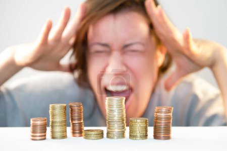 A man holds his head with his hands, screams with a grimace of horror, anxiety and helplessness, stress from lack of money in front of stacks of coins. Economic problems, financial crisis
