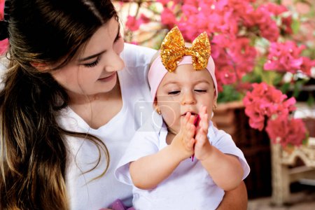 Eine junge Mutter lächelt, hält ein süßes kleines Mädchen im Alter von einem Jahr auf dem Arm, das Kind spielt interessiert und lustig mit einer Blume.
