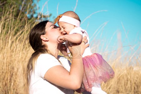 Die junge Mutter umarmt, küsst ein süßes lachendes Mädchen vor dem Hintergrund von rustikalem Feld und Himmel. Gefühle der Liebe, das Glück einer jungen Familie. Muttertag, Kindertag, Familientag
