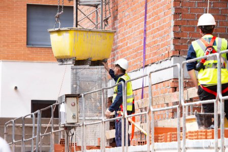 A worker in safety gear supervises the lifting of a container with a concrete mixture against the background of a brick wall, protective barriers at the height of a building under construction.