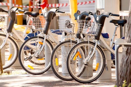 A group of rental electric bicycles parked at a docking station in an urban area with warning tape.