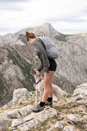 Téléchargez les photos : Plan vertical d'une jeune fille blanche vêtue de vêtements de montagne et d'un sac à dos gris caresse son chien.En arrière-plan se trouve une belle montagne voisine. Fonds d'écran pour la randonnée et l'escalade. - en image libre de droit