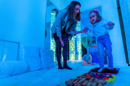 Therapist helping a young child living with cerebral palsy successfully improve her walking ability during therapy session in a sensory room, snoezelen. Texture walk, sensory exploration, motor skills.