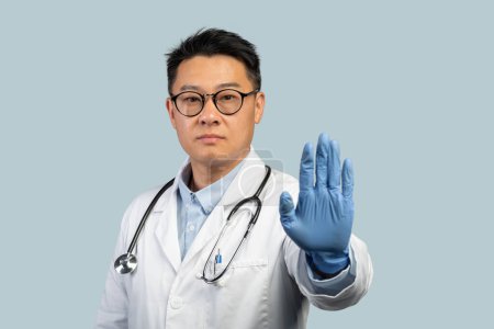 Foto de Confiado médico masculino chino de mediana edad en bata blanca, guantes de protección y gafas muestra señal de stop con la mano, aislado sobre fondo azul. Restricción y decir no a la enfermedad al aborto - Imagen libre de derechos