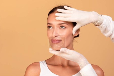 Kosmetikerin in Handschuhen überprüft vor Schönheitsbehandlung das Gesicht einer jungen Frau, berührt Stirn und Kinn auf beigem Hintergrund, freier Kopierraum