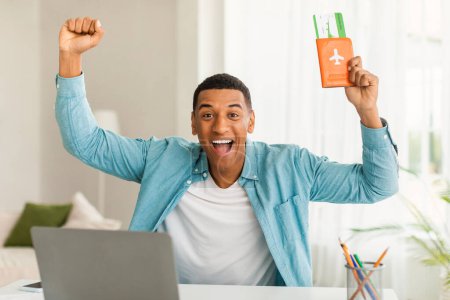 Photo for Laughing excited millennial black guy in casual rejoices to travel, victory, rises hands up with tickets and passport in living room office interior with pc. People emotions, vacation trip and travel - Royalty Free Image