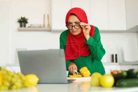 Foto de Señora musulmana madura cocinando mirando al ordenador portátil a través de anteojos preparando la cena a través de la receta en línea en la cocina moderna en interiores, usando Hijab. E-Learning And Healthy Food (en inglés). Enfoque selectivo - Imagen libre de derechos