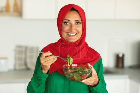 Foto de Alegre señora musulmana de mediana edad comiendo ensalada de verduras posando sosteniendo el tazón de fuente de pie en la cocina moderna en casa, sonriendo a la cámara. Nutrición saludable y pérdida de peso Recetas Concepto - Imagen libre de derechos
