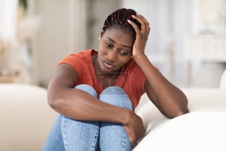 Portret przygnębionej młodej czarnej kobiety siedzącej na kanapie w domu, zdenerwowanej, zamyślonej, afroamerykańskiej kobiety z krzywą głową na dłoni i patrzącej w dal, cierpiącej na załamanie psychiczne lub sezonową depresję
