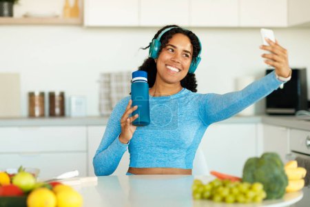 Téléchargez les photos : Femme afro-américaine tirant selfie et tenant bouteille portant des vêtements de sport, assis dans la cuisine après l'entraînement domestique. Concept de diétologie et nutrition - en image libre de droit