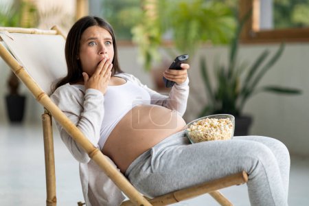 Foto de Contenidos de choque. Mujer embarazada asustada viendo la televisión y comiendo palomitas de maíz en casa, aterrorizada expectante hembra sosteniendo el mando a distancia y cubriendo la boca con la mano, reaccionando emocionalmente al programa de televisión - Imagen libre de derechos