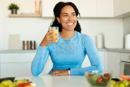 Photo for Positive black lady in sportswear sitting in kitchen and drinking fresh orange juice from glass before or after workout at home. Active healthy lifestyle, clean eating concept - Royalty Free Image