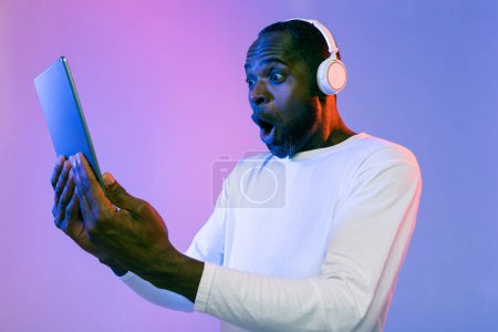 Téléchargez les photos : Émotionnel homme noir d'âge moyen en tenue décontractée avec casque sans fil regardant l'écran de la tablette numérique sur fond de studio néon, regarder la publicité cool en ligne, en utilisant une belle application divertissante - en image libre de droit