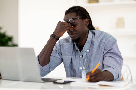 Photo for Tired Young Black Man Suffering Eye Strain While Working With Laptop At Desk, Stressed African American Male Massaging Nosebridge And Frowning, Having Migraine Or Eyes Fatigue, Free Space - Royalty Free Image