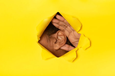 Téléchargez les photos : Une goutte d'eau. Femme afro-américaine méconnaissable mettant l'oreille au papier jaune déchiré écoutant à travers le trou en arrière-plan studio. Je t'entends, concept d'offre secrète. Collage - en image libre de droit