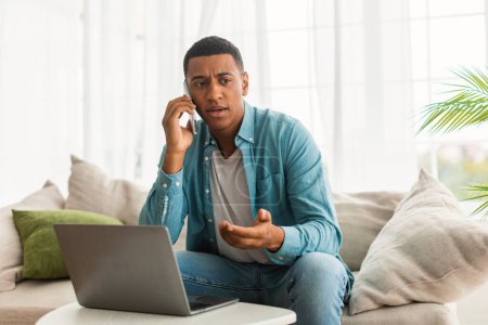 Téléchargez les photos : Malheureux sérieux occupé jeune afro-américain travail masculin avec ordinateur, appels par smartphone dans la lumière intérieur du salon. Résoudre les problèmes liés à la télécommande d'affaires à domicile, au soutien à la clientèle, à la pandémie covid-19 - en image libre de droit