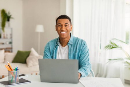 Téléchargez les photos : Souriant jeune homme d'affaires afro-américain ou gestionnaire utilise l'ordinateur pour étudier, travailler dans la salle de lumière moderne ou l'intérieur du bureau. Entreprise, freelance à distance, distance sociale à la maison covid-19 pandémie - en image libre de droit