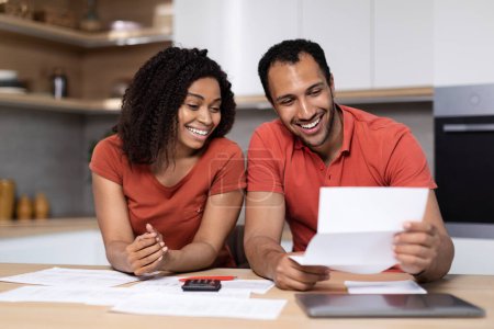 Happy young black lady in red t-shirt shows documents to guy with computer at kitchen interior. Great news, bill payment, home bookkeeping, taxes together, household chores, end of mortgage and credit