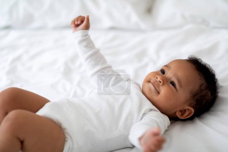 Photo for Closeup Portrait Of Cute Little African American Baby Lying On Bed, Small Adorable Black Infant Boy Or Girl Wearing Bodysuit Resting On White Linens In Bedroom, Spreading His Arms And Smiling - Royalty Free Image