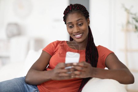 Photo for Beautiful Young Black Woman Messaging On Smartphone While Relaxing At Home, Smiling African American Female Sitting On Couch And Using Mobile Phone For Online Coomunication, Copy Space - Royalty Free Image