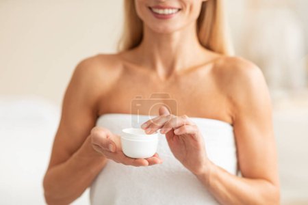 Photo for Unrecognizable mature woman holding opened jar with moisturising cream, female wearing white towel and making beauty skincare routine at home, cropped - Royalty Free Image