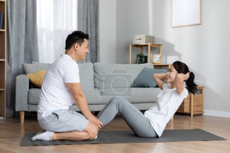 Seitenansicht athletischer koreanischer Mann und Frau in weißer Sportbekleidung, die zusammen trainieren, an der Bauchmuskulatur arbeiten, sich gegenseitig ermutigen und unterstützen, während sie zu Hause trainieren, Raum kopieren