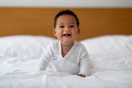 Photo for Adorable Cute Black Baby Lying On Tummy While Relaxing On Bed At Home, Closeup Portrait Of Smiling Little Infant African American Boy Or Girl Resting On White Linens In Bedroom, Copy Space - Royalty Free Image