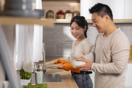 Téléchargez les photos : Heureux conjoints asiatiques faisant des corvées ensemble, belle femme lavant la vaisselle et aimant mari séchant les assiettes, ayant la conversation et souriant, vue de côté, espace de copie - en image libre de droit