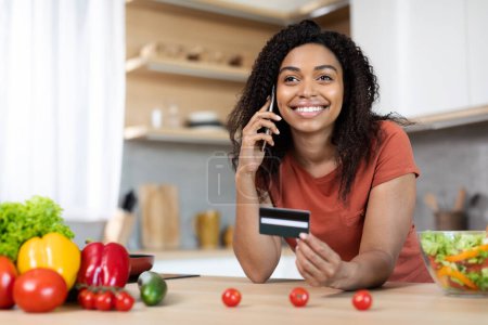 Foto de Feliz joven hembra negra en camiseta roja llamando por teléfono, mostrando la tarjeta de crédito en la mesa con verduras orgánicas en el interior de la cocina moderna. Pedido de comestibles en casa, compras en línea, venta y servicio - Imagen libre de derechos