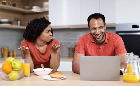 Traurige unzufriedene junge schwarze Frau nimmt es übel und beschimpft lächelnden Ehemann mit PC in der Küche. Verrat in sozialen Netzwerken, Eifersucht, Glücksspiel, Spielsucht und Beziehungsprobleme