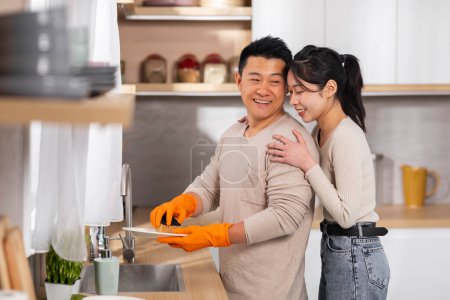 Photo for Positive attractive middle aged chinese man in colorful gloves washing dishes, helping his wife with chores at home, thankful young lady hugging her boyfriend from behind, copy space - Royalty Free Image