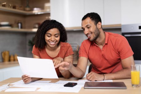 Cheerful young black female in red t-shirt shows documents to man with pc, check accounts use banking at kitchen interior. Bills pay together, bookkeeping, taxes, household chores, mortgage and profit
