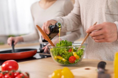 Unrecognizable spouses cooking dinner together at home, cropped of wife and husband preparing healthy meal, making vegetable salad, adding olive oil, copy space. Nutrition, healthy diet concept