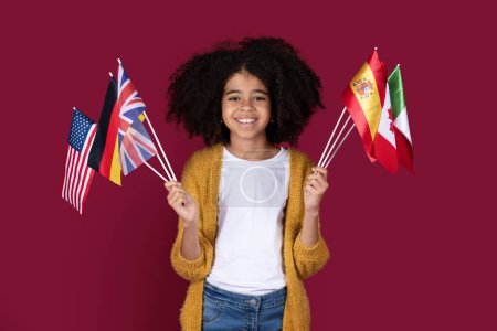 Téléchargez les photos : Adorable fille noire aux cheveux touffus en tenue décontractée écolière tenant un tas de drapeaux de divers pays et souriant joyeusement sur fond de studio bordeaux. Éducation des enfants à l'étranger concept - en image libre de droit