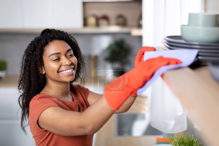 Photo for Cheerful pretty young black woman in red t-shirt and rubber gloves dusts off from wooden shelf in kitchen interior, copy space. Household chores, perfectionism, cleanliness, hygiene and health care - Royalty Free Image