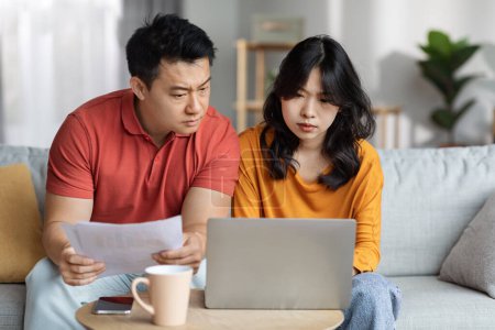 Photo for Upset asian couple checking bills, sitting on couch, reading documents, using laptop, unhappy chinese man and woman holding papers, counting monthly spendings, copy space. Family budget concept - Royalty Free Image