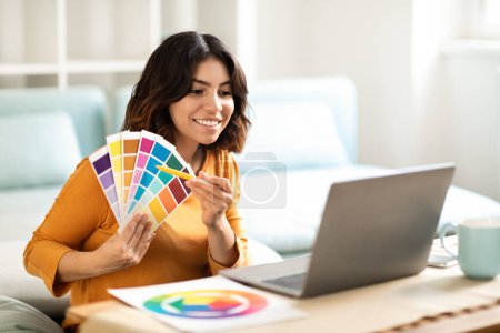 Photo for Young Arab Interior Designer Lady Showing Swatches To Client Via Teleconference On Laptop, Smiling Middle Eastern Woman Demonstrating Color Examples During Video Call On Computer, Free Space - Royalty Free Image